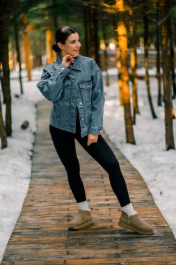 Genç bir kadın güneşli bir kış günü, günlük kıyafetleriyle, karlı bir ormanda, tahta bir patikada yürüyor.