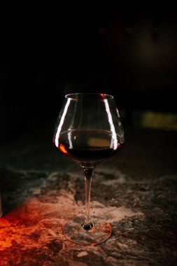 Koyu yüzeyde, kırmızı şarap kadehinin yanında ince yansımalar ve sıcak ışık efektleri var.