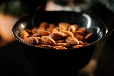 Taze kavrulmuş bademler, siyah bir kâsede altın-kahverengi rengini vurgulayan koyu bir arka plana karşı servis edilir.