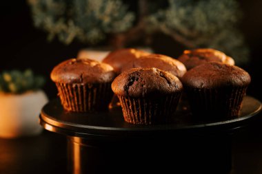 Loş bir mutfak ortamında yuvarlak siyah bir tabakta taze pişmiş çikolatalı kekler.