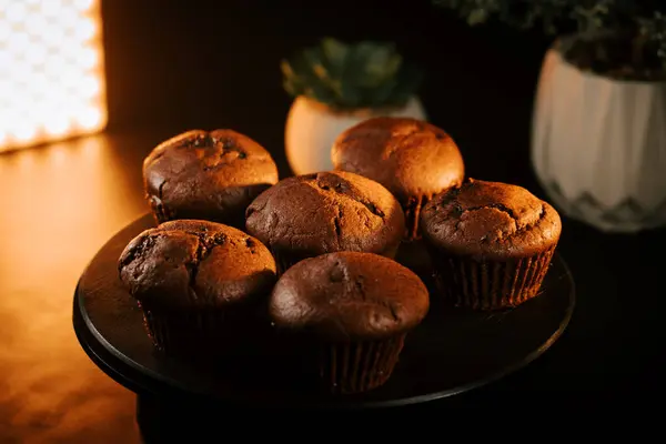 Loş bir mutfak ortamında yuvarlak siyah bir tabakta taze pişmiş çikolatalı kekler.
