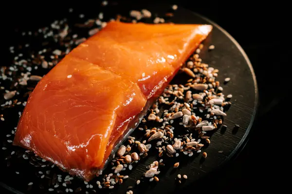 Tohumlarla çevrili siyah servis tahtasındaki taze organik alabalık filetosu.