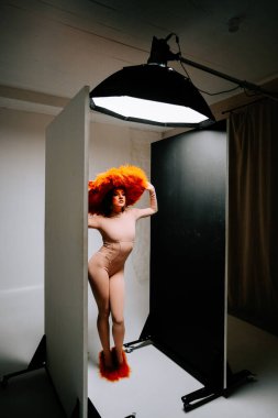 Model, yaratıcı bir moda çekimi sırasında minimalist bir stüdyoda canlı turuncu şapkayla dramatik bir şekilde poz verir.
