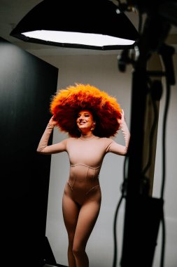 Model, yaratıcı bir moda çekimi sırasında minimalist bir stüdyoda canlı turuncu şapkayla dramatik bir şekilde poz verir.