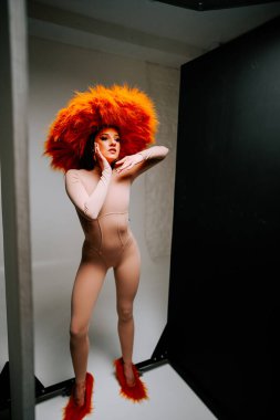 Model, yaratıcı bir moda çekimi sırasında minimalist bir stüdyoda canlı turuncu şapkayla dramatik bir şekilde poz verir.