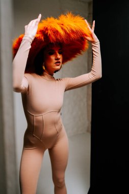 Model, yaratıcı bir moda çekimi sırasında minimalist bir stüdyoda canlı turuncu şapkayla dramatik bir şekilde poz verir.