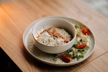 Üzerine kızarmış pastırma ve taze salatayla servis edilen parmesan soslu yulaf ezmesi.