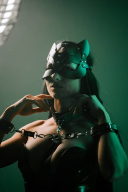 Kedi maskeli ve kelepçeli kadın karanlık bir ortamda neon ışıkların altında poz veriyor.