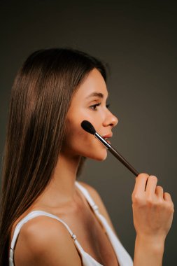 Kadın içeride tarafsız bir arkaplanda poz verirken fırçayla makyaj yapıyor.