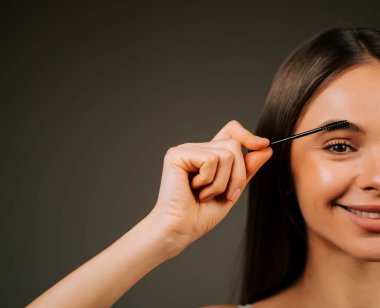 Kadın kaşlarını tımar etmek için kaş fırçası kullanıyor. Gülümsüyor.
