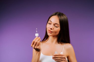 Kadın, mor arka planda pipetle yüz serumu uyguluyor.