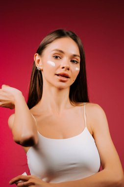 Cilt bakımı rutini. Stüdyoda pembe arka planı olan bir kadın krem sürüyor.