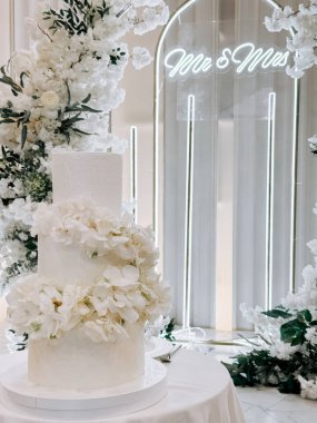 Beyaz çiçeklerle süslenmiş zarif bir düğün pastası ve lüks bir yerde ışıklandırılmış bir fon.