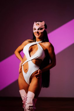 Stüdyoda seksi kıyafetler ve neşeli ifadeli pembe kedi maskesi giyen bir bayan dansçı poz veriyor.
