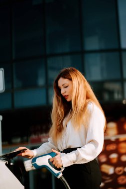 Şehir merkezinde güneşli bir günde şarj istasyonunda bir kadın elektrikli araba yüklüyor.