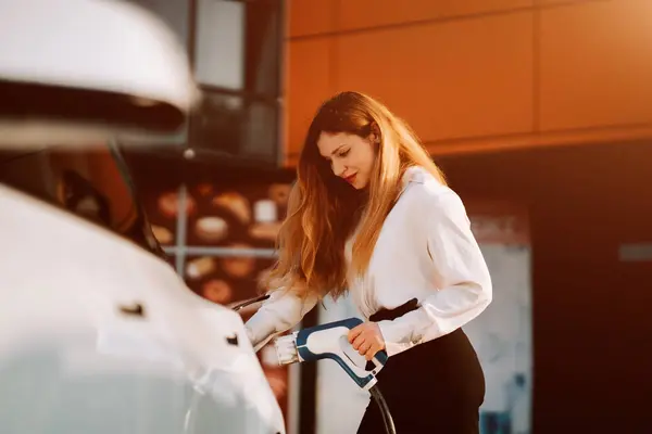 Gündüz vakti modern bir şehir ortamında bir kadın elektrikli araba şarj ediyor.