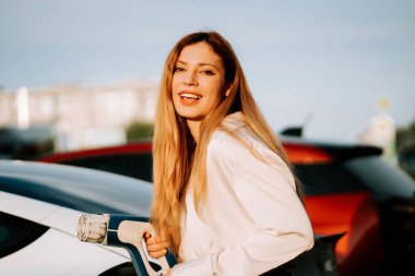 Kızıl saçlı genç bir kadın güneşli bir günde alışveriş merkezinin yanındaki istasyonda elektrikli araç kullanıyor.