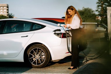 Kızıl saçlı kadın, alışveriş merkezinin yanındaki istasyonda elektrikli arabasını şarj ediyor.