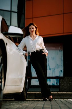 Kızıl saçlı genç bir kadın gün ışığında alışveriş merkezinin yakınındaki bir istasyonda elektrikli arabasını şarj ediyor.