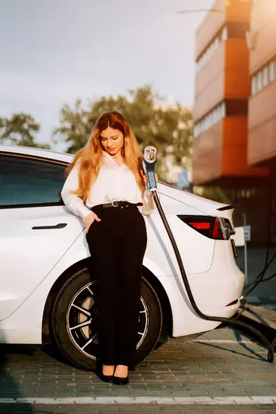 Kızıl saçlı genç bir kadın gün batımında alışveriş merkezinde elektrikli aracını şarj ediyor.