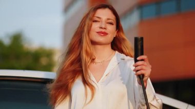 Şehir ortamında gün batımında genç bir kadın elektrikli araba şarj ediyor.