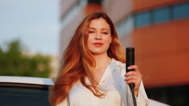 Genç kadın gün batımında şarj istasyonunda elektrikli araç yüklüyor.