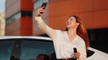 Genç bir kadın dışarıda selfie çekerken elektrikli arabasını şarj etmekten hoşlanıyor.
