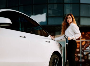 Kadın gün batımında park yerinde elektrikli arabayı sakin bir arka planla şarj ediyor.