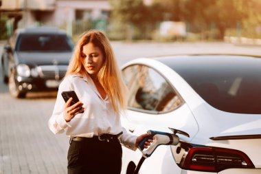 Kadın gün batımında otoparkta telefonu kontrol ederken elektrikli arabayı şarj ediyor.