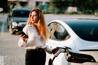 Gün batımında kadın elinde akıllı telefonla otoparkta elektrikli arabayı şarj ediyor.