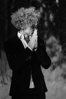 Siyah takım elbiseli kıvırcık saçlı, karlı bir ormanda tefekkür eden biri.