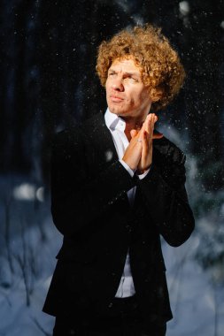 Siyah takım elbiseli adam, kış güneşinde umut dolu ifadesiyle karlı ağaçların arasında duruyor.