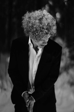 Takım elbiseli, kıvırcık saçlı bir adam ormanda dalgın dalgın poz veriyor.