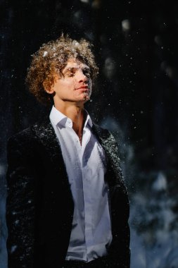 Karanlık bir ormanda kar yağışı seven genç bir adam, resmi giyinmiş ve doğayı kucaklıyor.