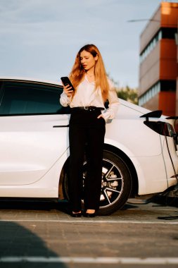 Kızıl saçlı genç bir kadın gün batımı şarj istasyonunda elektrikli arabasını şarj ediyor.