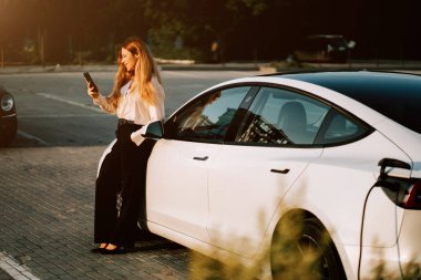 Kızıl saçlı genç kadın gün batımında şehir şarj istasyonunun yakınında elektrikli araba yüklüyor.
