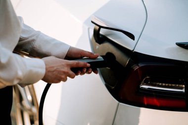 Kızıl saçlı genç bir kadın gün batımında şarj istasyonunun yanında elektrikli araba yüklüyor.