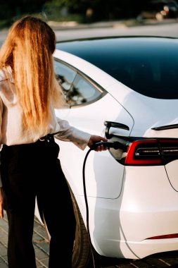 Kızıl saçlı genç bir kadın şarj istasyonunda günbatımında elektrikli araba yüklüyor.