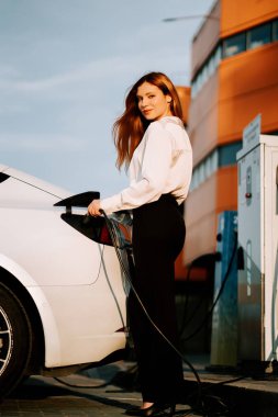 Kızıl saçlı genç kadın gün batımında şehir şarj istasyonunun yakınında elektrikli araç yüklüyor.