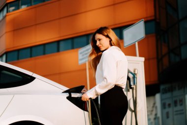 Kızıl saçlı genç kadın gün batımında şarj istasyonunda elektrikli araba şarj ediyor.