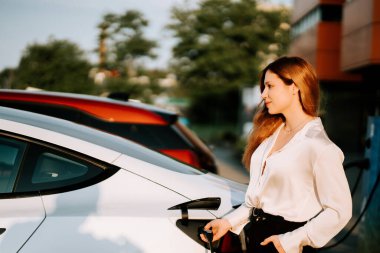 Kızıl saçlı genç kadın gün batımında şehir şarj istasyonunun yakınında elektrikli araba yüklüyor.