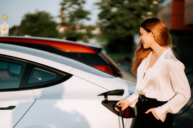 Kızıl saçlı genç bir kadın sakin bir ortamda gün batımında elektrikli arabasını şarj ediyor.