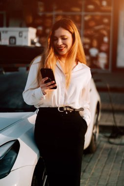 Kızıl saçlı genç bir kadın gün batımında akıllı telefonunu kullanırken elektrikli arabayı şarj ediyor.