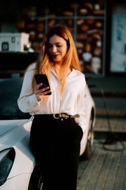 Kızıl saçlı genç bir kadın gün batımında telefonunu kontrol ederken elektrikli arabasını şarj ediyor.