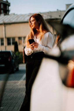 Kızıl saçlı genç bir kadın şehir merkezinde günbatımında elektrikli araba kullanıyor.