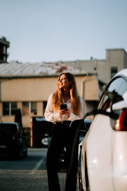 Kızıl saçlı genç bir kadın gün batımında şarj istasyonunun yakınında elektrikli araba kullanıyor.