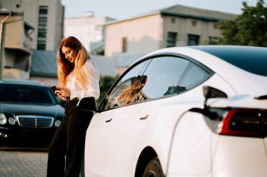 Kızıl saçlı genç bir kadın günbatımı şarj istasyonunda elektrikli arabaya güç veriyor.