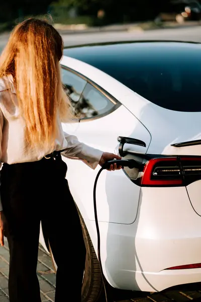 Kızıl saçlı genç bir kadın şarj istasyonunda günbatımında elektrikli araba yüklüyor.