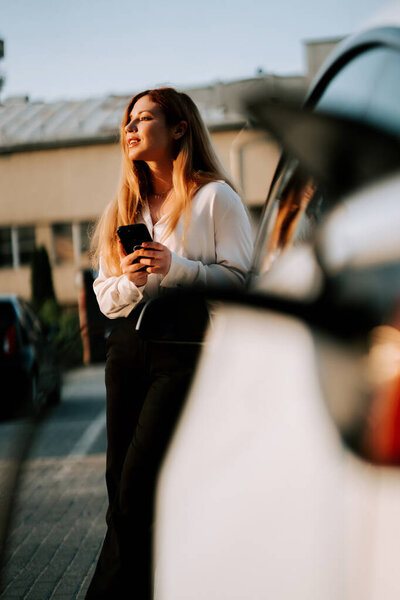 Молодая женщина с рыжими волосами заряжает электрический автомобиль на закате на зарядной станции