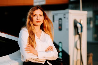 Kızıl saçlı bir iş kadını gün batımında elektrikli arabayı şarj ediyor.